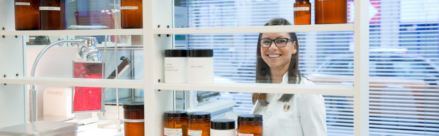 Durch die Regale im Labor, auf denen braune Flaschen mit Tinkturen und Lösungen stehen sieht man eine freundlich lächelnde Apothekerin bei ihrer Arbeit